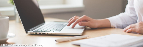 Frau arbeitet am Computer, neben Ihr auf dem Schreibtisch liegen ein Stift und ein Notitzbuchh